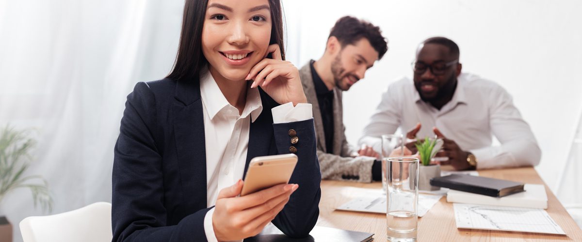Portrait of beautiful asian businesswoman sitting in office and joyfully looking in camera with cellphone in hands and multinational business partners on background
