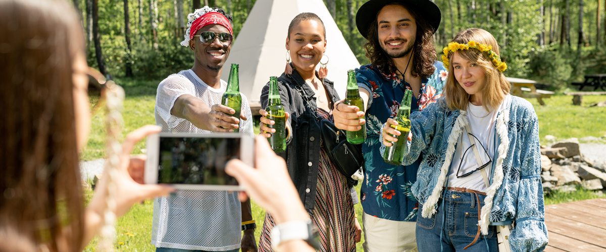 Group of positive young multi-ethnic people in hippie outfits posing with beer bottles for photo at countryside festival